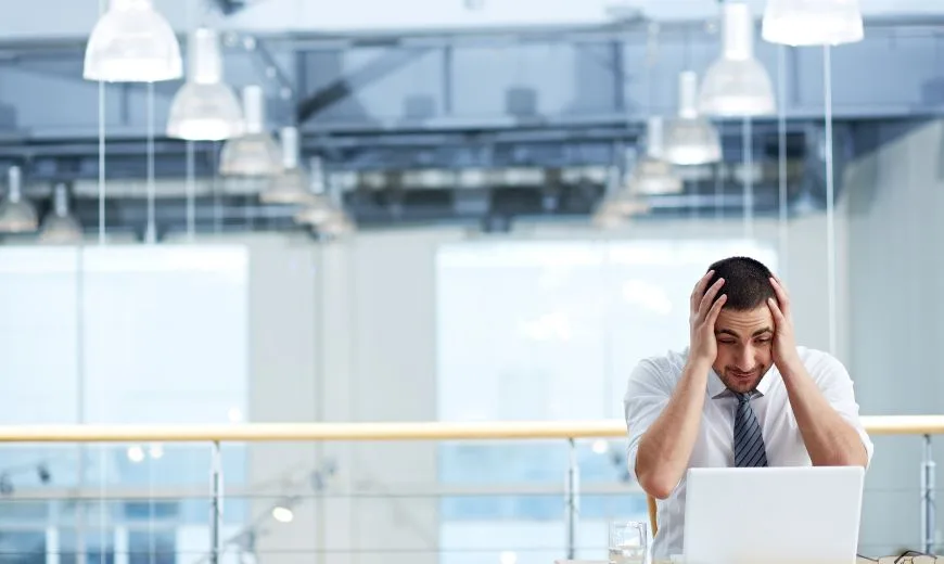 Hombre frustrado agarrándose la cabeza mientras trabaja con una computadora portátil en una oficina luminosa.